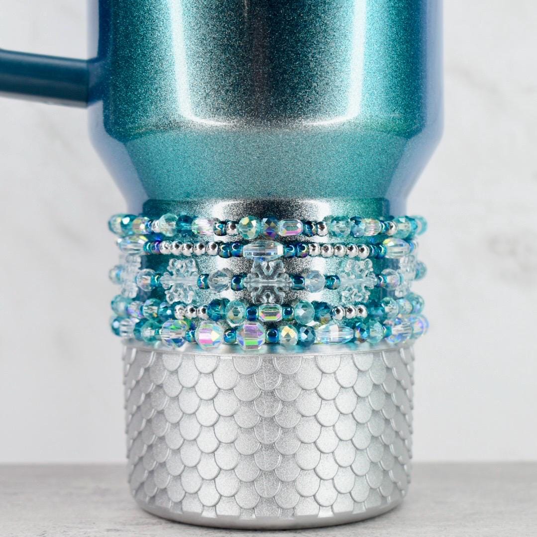 A teal and silver tumbler with a beaded accent. The upper portion of the tumbler is teal with a glittery finish. The middle section features multiple rows of blue and clear beads. The bottom has a silver, scale-like design.