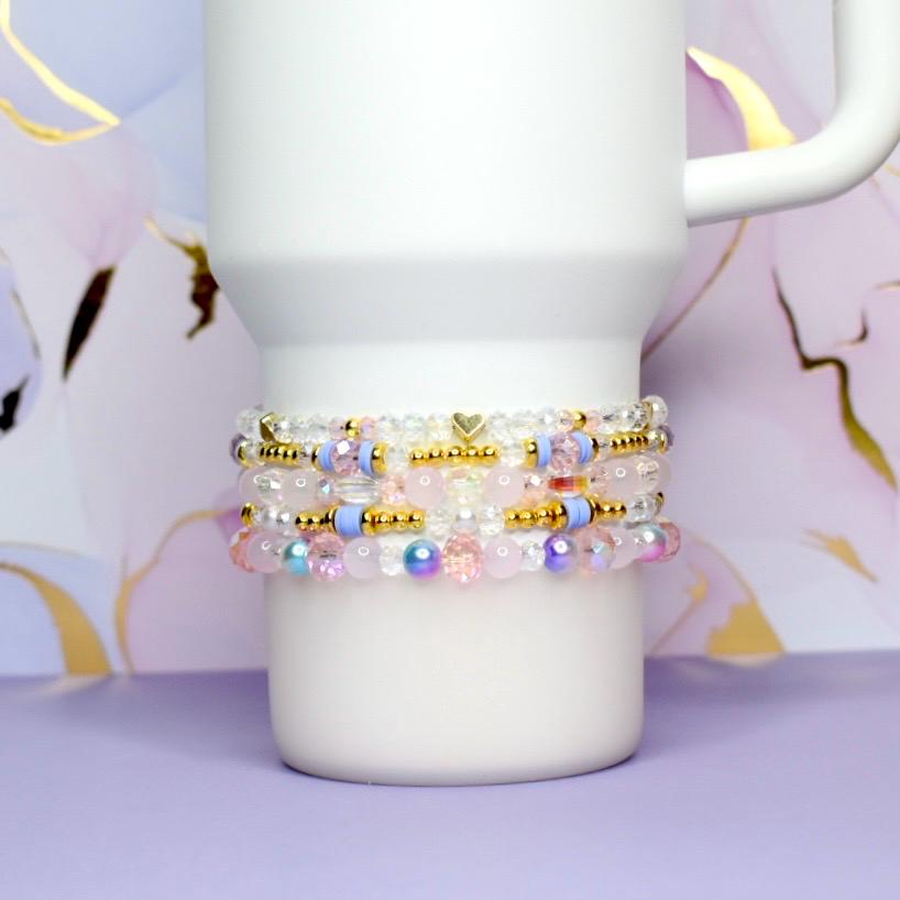 Stack of beaded bracelets in shades of pink, white, gold, and blue. The bracelets are made with various beads, including clear, iridescent, and gold-colored beads. A white mug is in the background, resting on a purple surface.