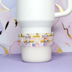 Stack of beaded bracelets in shades of pink, white, gold, and blue. The bracelets are made with various beads, including clear, iridescent, and gold-colored beads. A white mug is in the background, resting on a purple surface.