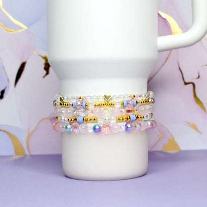 Stack of beaded bracelets in shades of pink, white, gold, and blue. The bracelets are made with various beads, including clear, iridescent, and gold-colored beads. A white mug is in the background, resting on a purple surface.