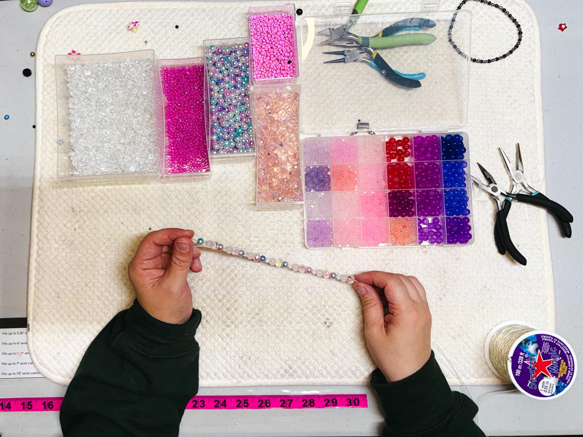 A craft setup with various beads in clear containers, a divided organizer with colorful beads, and jewelry-making tools. A partially completed beaded bracelet is held by hands. A measuring tape is visible.