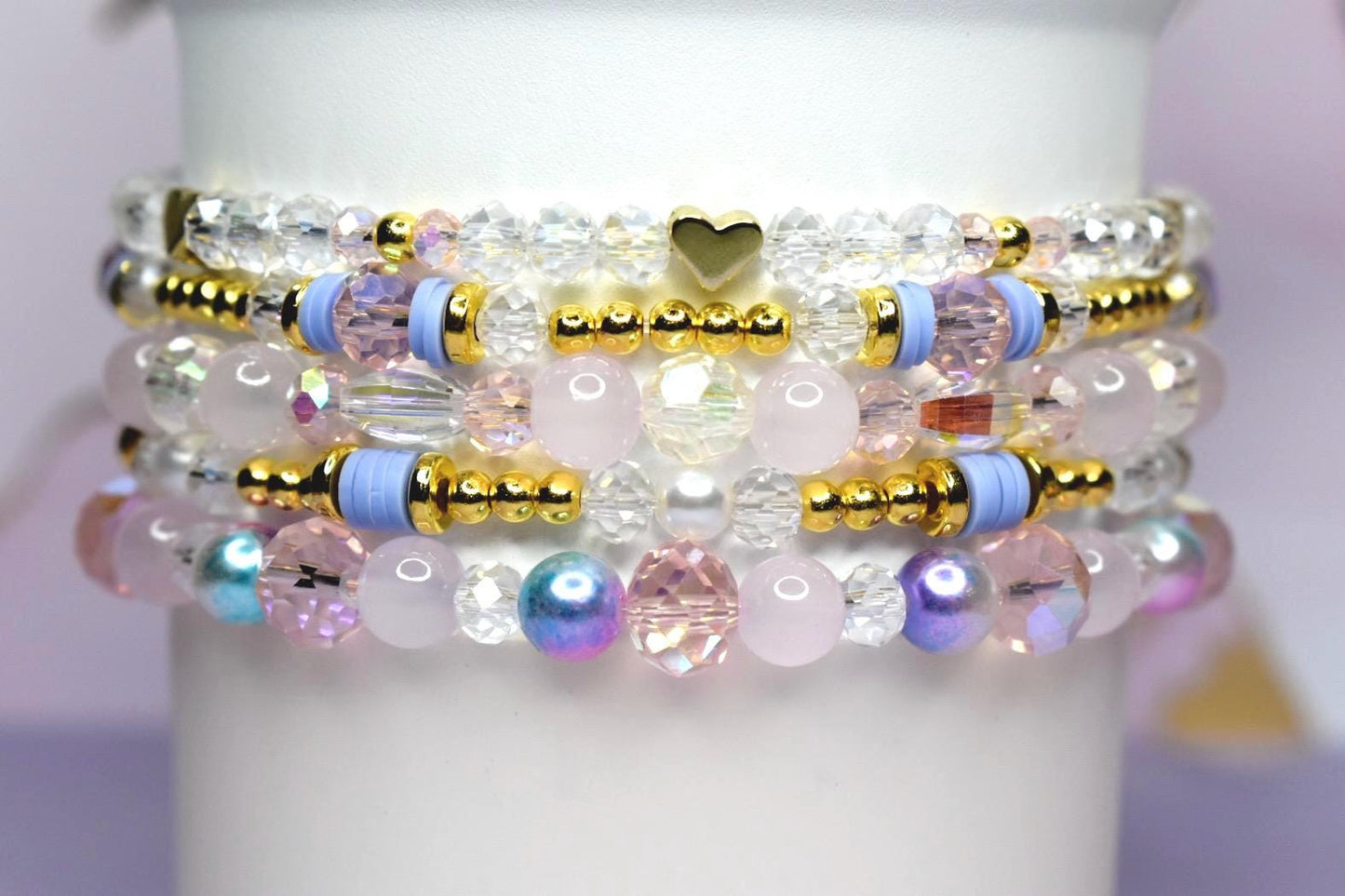 Stack of beaded bracelets in shades of pink, white, and iridescent colors. The bracelets feature gold accents, clear crystal beads, and a small gold heart charm. The bracelets are stacked on a white cylindrical surface.