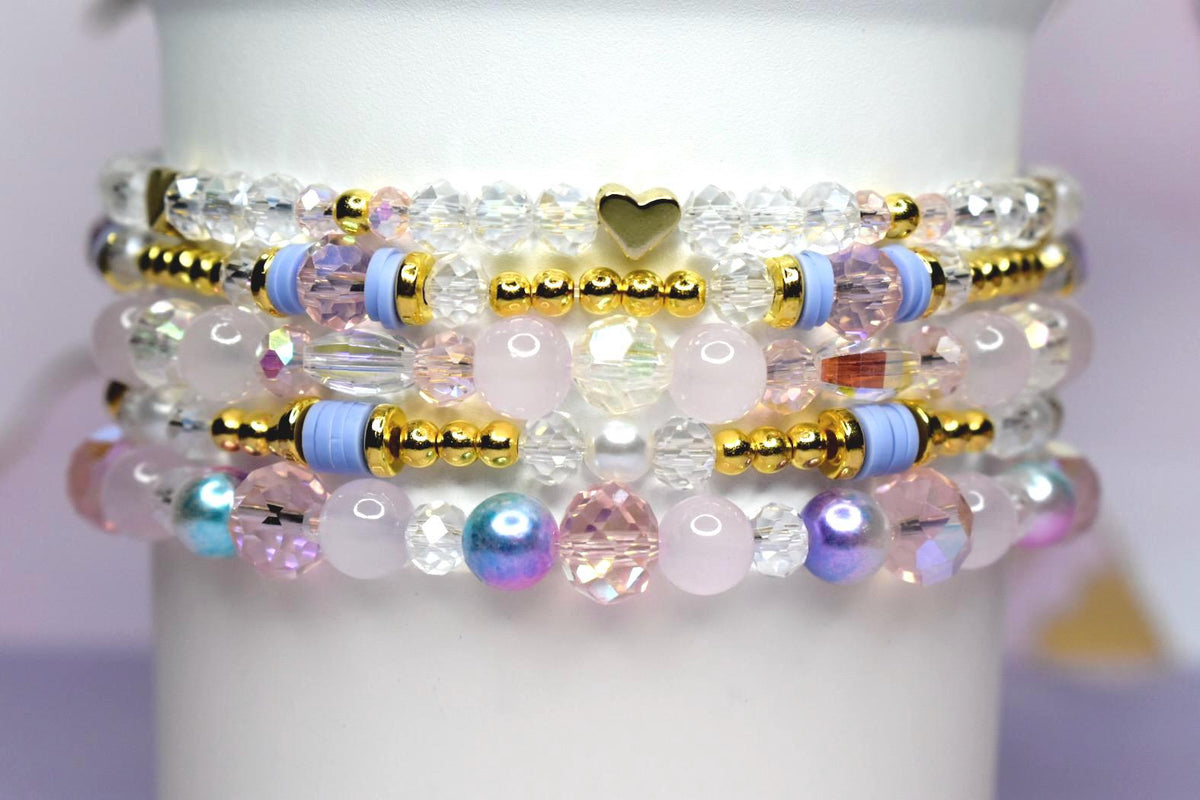 Stack of beaded bracelets in shades of pink, white, and iridescent colors. The bracelets feature gold accents, clear crystal beads, and a small gold heart charm. The bracelets are stacked on a white cylindrical surface.