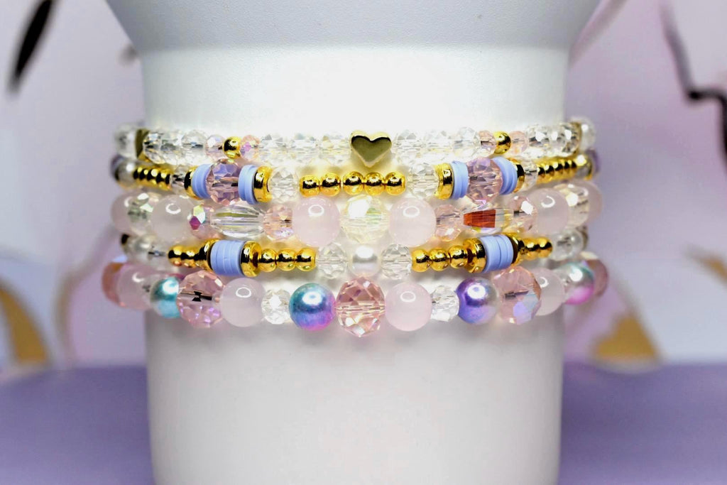 Stack of beaded bracelets in shades of pink, gold, and clear. The bracelets feature a variety of beads, including clear crystals, gold accents, and pink and iridescent beads. A small gold heart charm is visible.