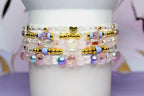 Stack of beaded bracelets in shades of pink, gold, and clear. The bracelets feature a variety of beads, including clear crystals, gold accents, and pink and iridescent beads. A small gold heart charm is visible.