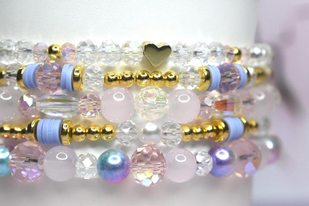 Stack of beaded bracelets in shades of pink, purple, and clear. The bracelets feature gold accents, clear crystal beads, and a gold heart charm. Some bracelets have light blue spacers.