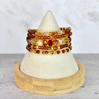 A stack of beaded bracelets in shades of amber, gold, and brown. The bracelets are displayed on a white cone-shaped stand with a wooden base. The beads vary in size and shape, creating a layered effect.