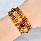 A collection of beaded bracelets in shades of amber, gold, and brown. The bracelets feature a variety of bead sizes and shapes, including round, faceted, and pearl-like beads. The bracelets are stacked together, creating a layered look.