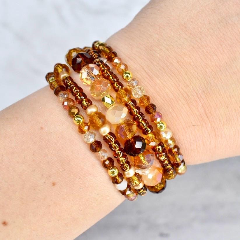 A collection of beaded bracelets in shades of amber, gold, and brown. The bracelets feature a variety of bead sizes and shapes, including round, faceted, and pearl-like beads. The bracelets are stacked together, creating a layered look.