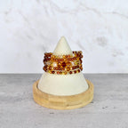 A stack of beaded bracelets in shades of amber, gold, and brown. The bracelets are displayed on a white cone-shaped stand with a wooden base. The beads vary in size and texture, creating a layered effect.