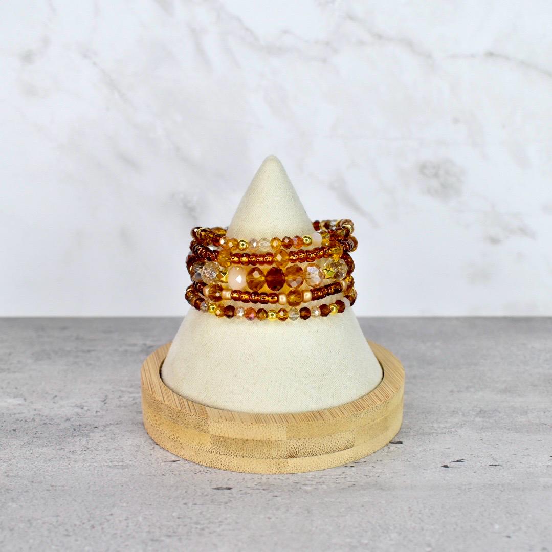 A stack of beaded bracelets in shades of amber, gold, and brown. The bracelets are displayed on a white cone-shaped stand with a wooden base. The beads vary in size and texture, creating a layered effect.