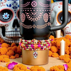 A black insulated tumbler with a handle, adorned with pink floral and skull designs. A stack of colorful beaded bracelets in shades of pink, orange, and gold wraps around the base. A silver skull charm with red accents hangs from the bracelets.