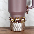 A mauve tumbler adorned with multiple beaded bracelets. The bracelets feature a mix of pink, purple, and gold beads. The tumbler has a textured, scale-like design at the base.