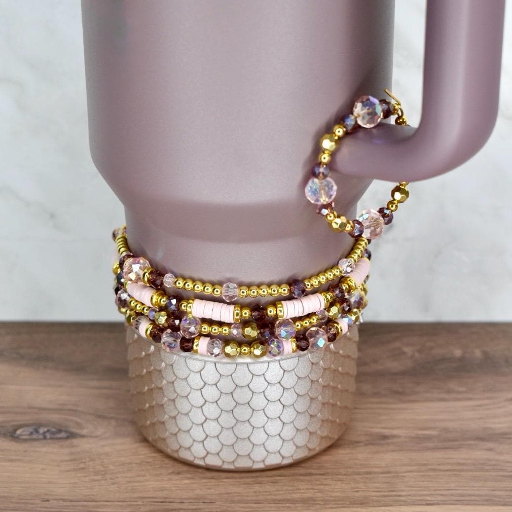 A collection of beaded bracelets in shades of pink, gold, and purple. The bracelets are stacked on a light purple tumbler with a gold, textured base. One bracelet is draped over the handle. The bracelets feature various bead sizes and shapes.