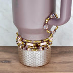 A collection of beaded bracelets in shades of pink, gold, and purple. The bracelets are stacked on a light purple tumbler with a gold, textured base. One bracelet is draped over the handle. The bracelets feature various bead sizes and shapes.