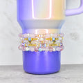 Stack of beaded bracelets in shades of pink, white, and gold, with iridescent and clear beads. The bracelets are stacked on a purple and iridescent tumbler. The bracelets have gold heart and blue accents.