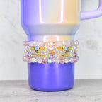 Stack of beaded bracelets in shades of pink, white, and gold, with iridescent and clear beads. The bracelets are stacked on a purple and iridescent tumbler. The bracelets have gold heart and blue accents.