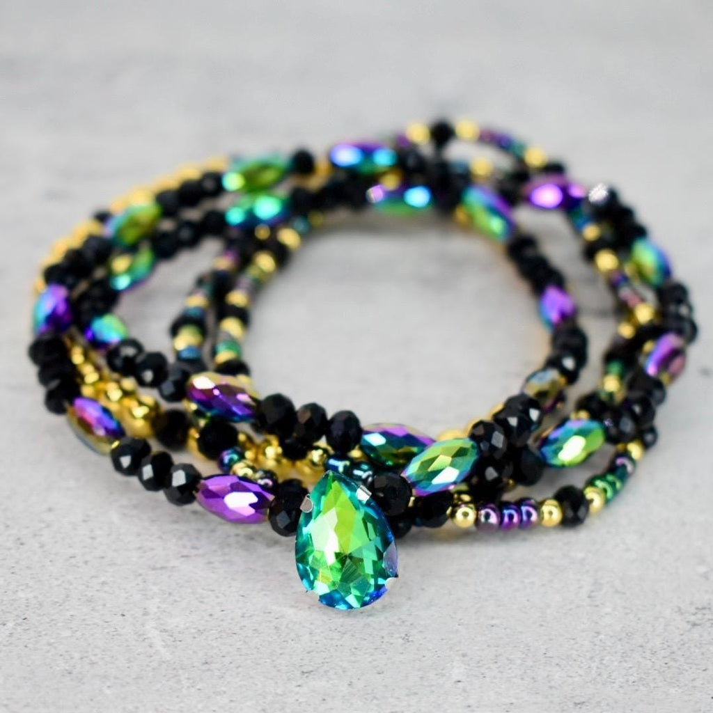 A multi-strand beaded bracelet featuring black, gold, and iridescent beads. A large, teardrop-shaped, green and blue crystal pendant hangs from the center of the bracelet. The bracelet is displayed on a light gray surface.