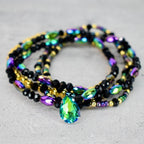 A multi-strand beaded bracelet featuring black, gold, and iridescent beads. A large, teardrop-shaped, green and blue crystal pendant hangs from the center of the bracelet. The bracelet is displayed on a light gray surface.