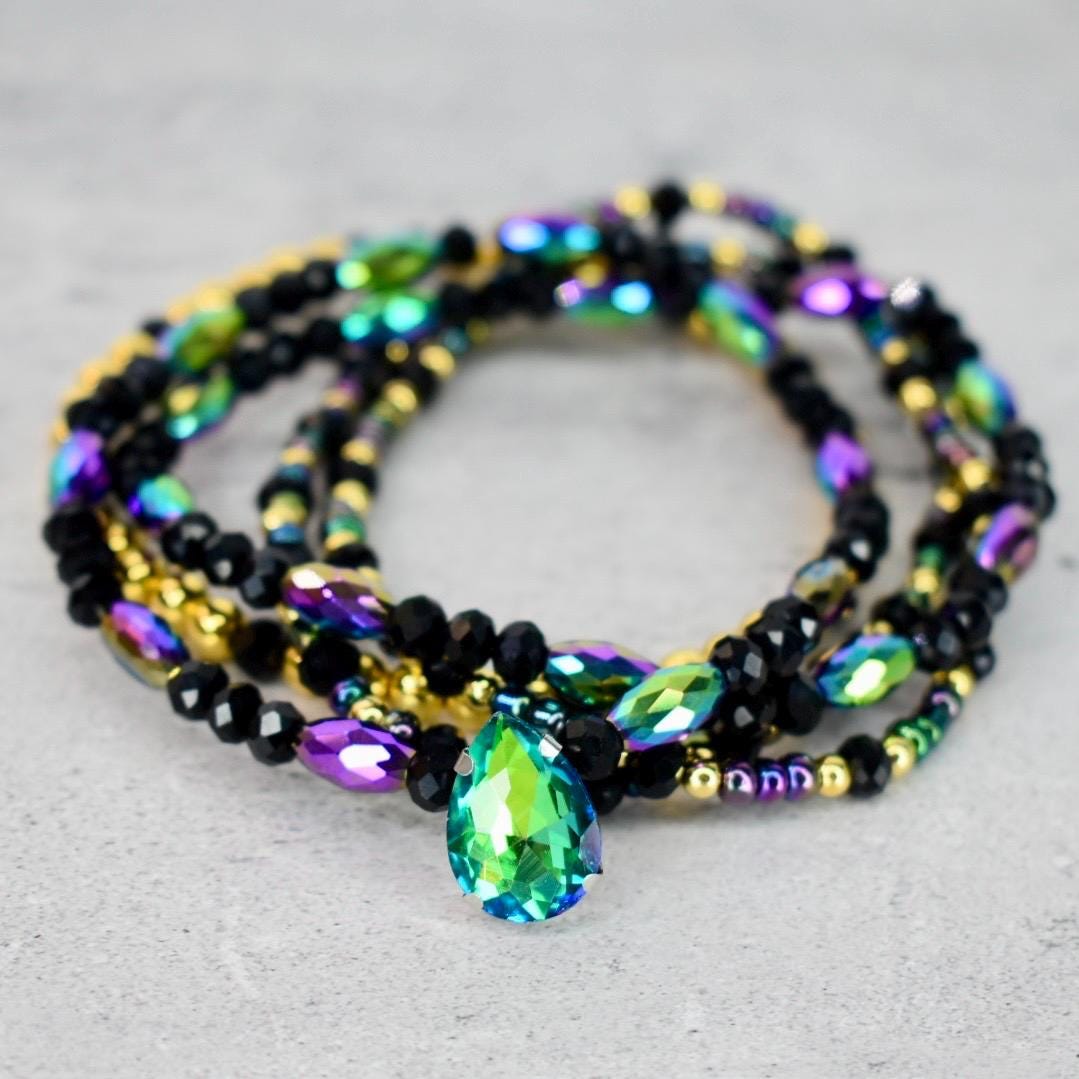 A multi-strand beaded bracelet featuring black, gold, and iridescent beads. A large, teardrop-shaped, green and blue crystal pendant hangs from the center of the bracelet. The bracelet is displayed on a light gray surface.