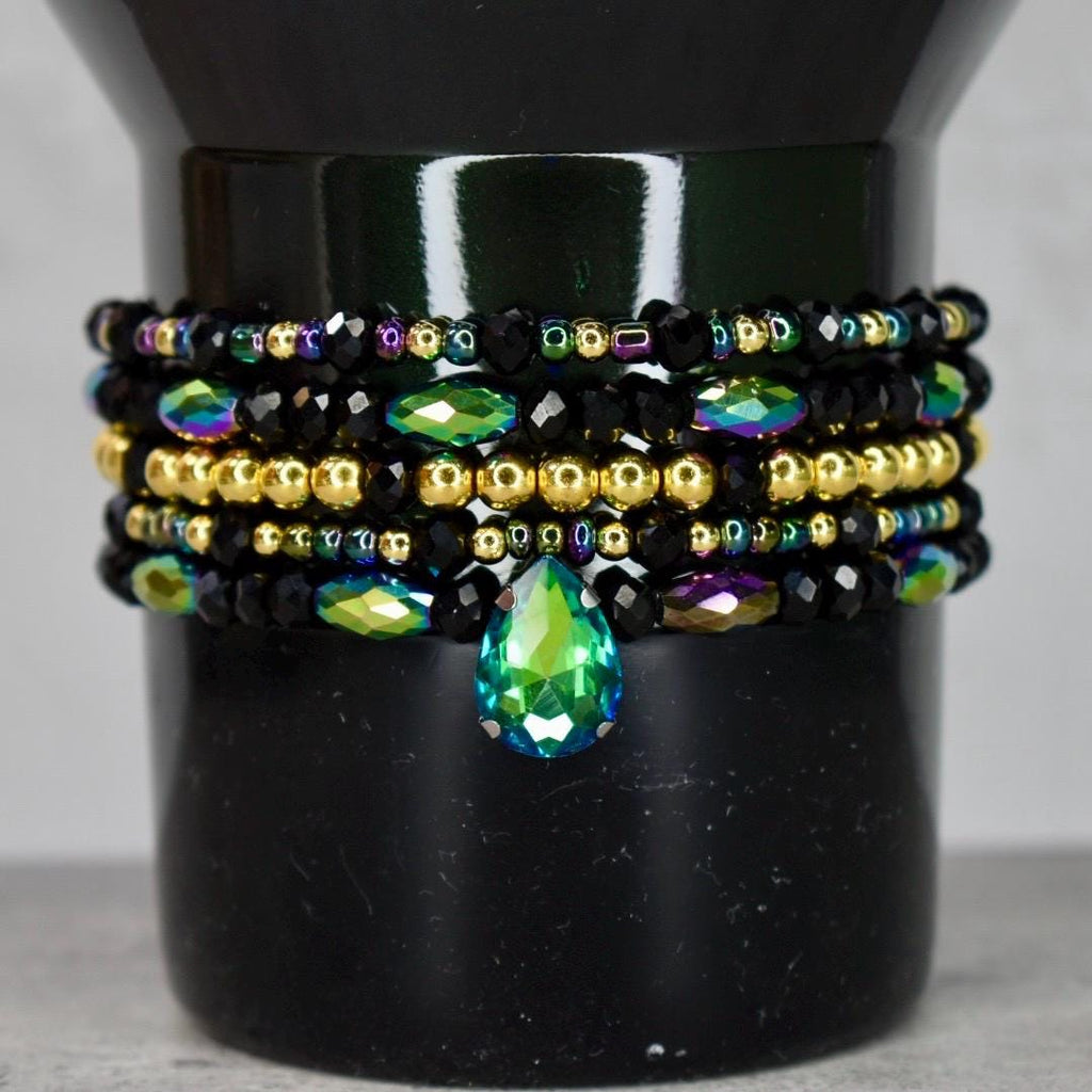 Stacked bracelets with black, gold, and iridescent beads. A large, teardrop-shaped green gem hangs from the bottom bracelet. The bracelets are displayed on the Elphaba For Good Stanley tumbler.
