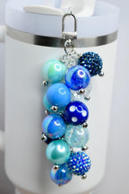 A blue beaded keychain with various shades of blue beads, including iridescent, solid, and textured spheres. The keychain is attached to a silver clasp and hangs in front of a white mug.