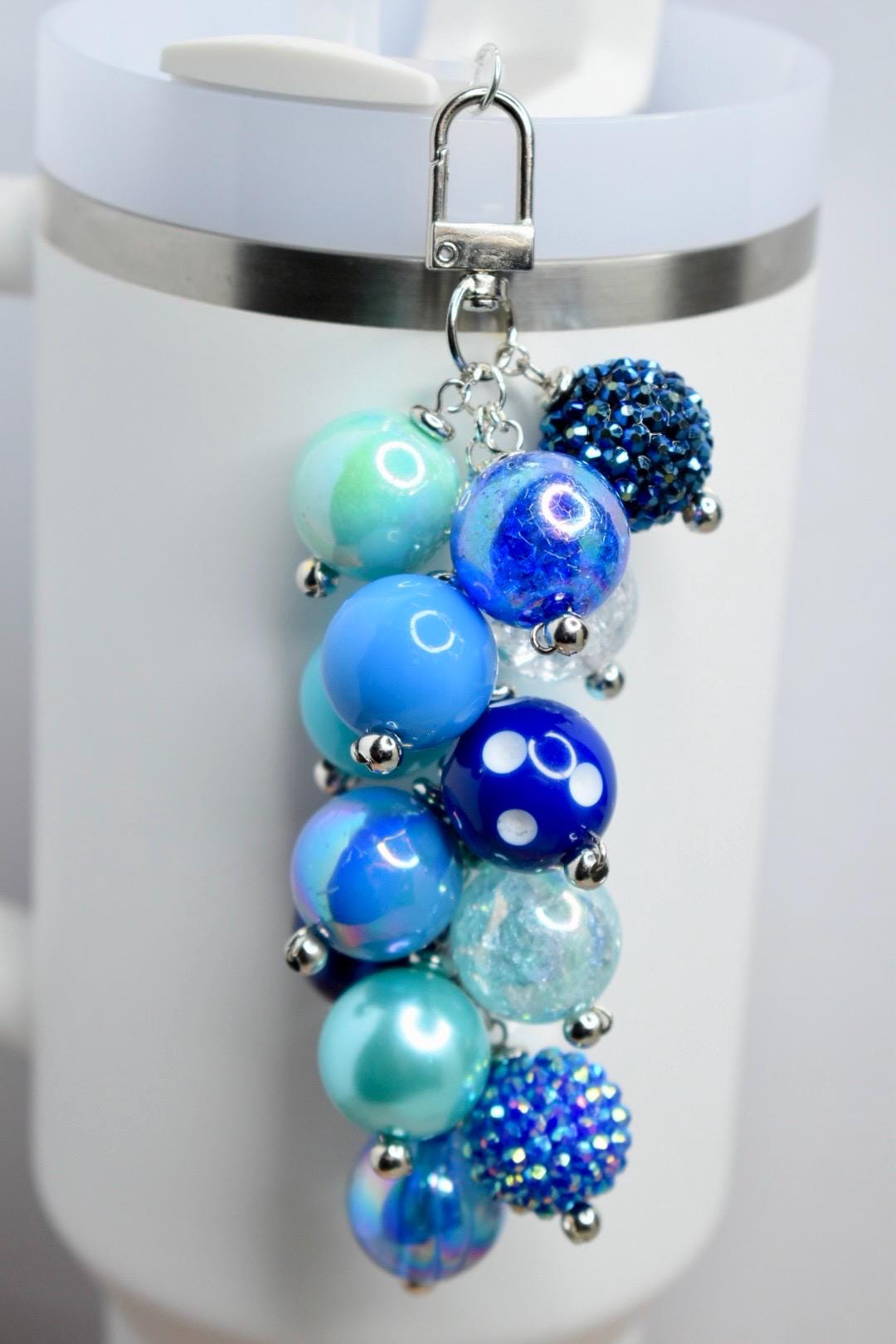 A blue beaded keychain with various shades of blue beads, including iridescent, solid, and textured spheres. The keychain is attached to a silver clasp and hangs in front of a white mug.