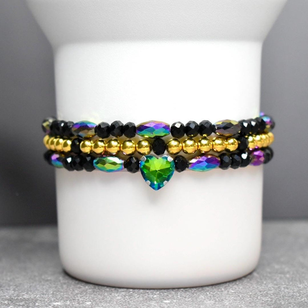 Stacked beaded bracelets in black, gold, and iridescent colors. The top and bottom bracelets feature black beads and iridescent accents. A gold beaded bracelet sits in the middle. A green heart-shaped charm hangs from the bottom bracelet.