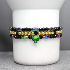 Stacked beaded bracelets in black, gold, and iridescent colors. The top and bottom bracelets feature black beads and iridescent accents. A gold beaded bracelet sits in the middle. A green heart-shaped charm hangs from the bottom bracelet.