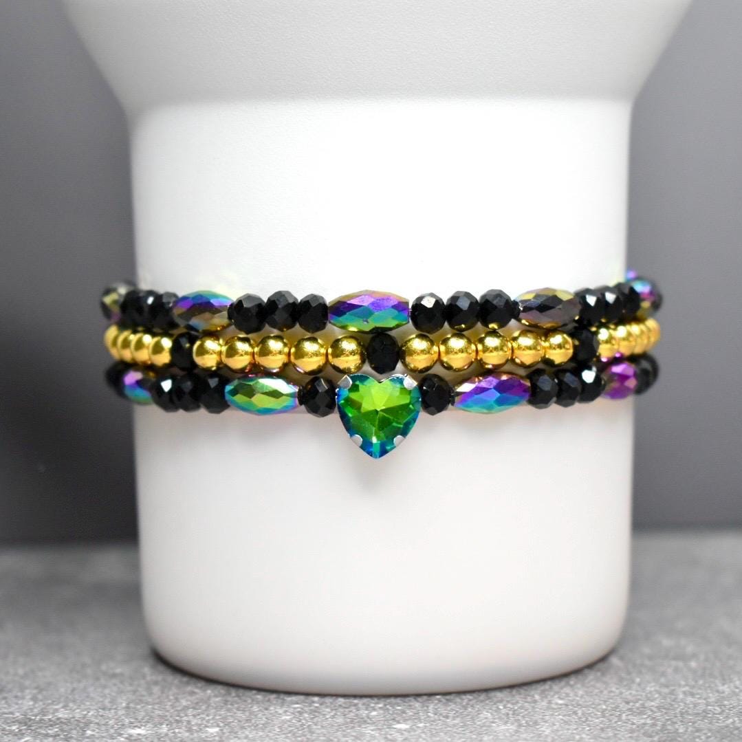 Stacked beaded bracelets in black, gold, and iridescent colors. The top and bottom bracelets feature black beads and iridescent accents. A gold beaded bracelet sits in the middle. A green heart-shaped charm hangs from the bottom bracelet.