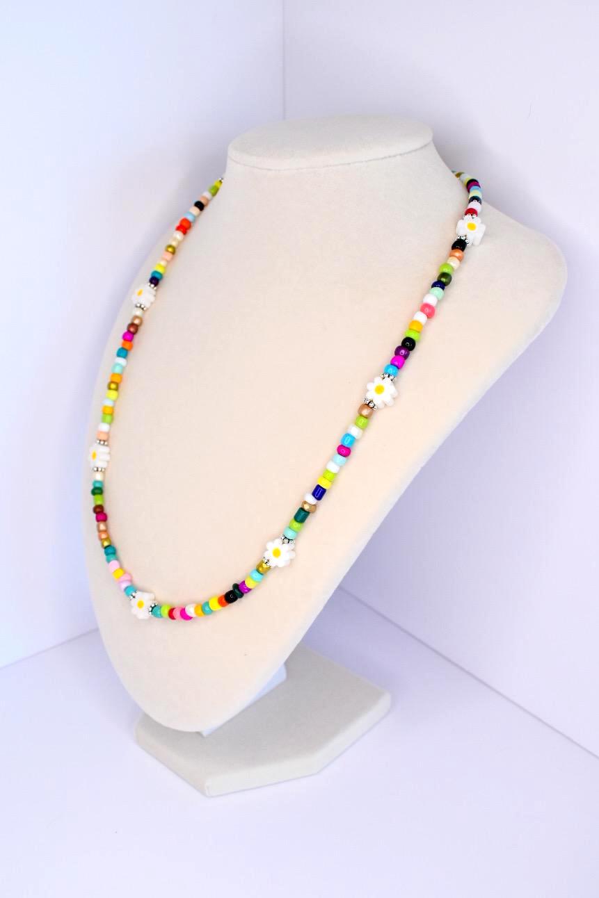 A colorful beaded necklace with daisy flower accents. The necklace features a variety of small, round beads in shades of red, yellow, green, blue, and black. White daisy-shaped beads are interspersed throughout the necklace. The necklace is displayed on a white mannequin.