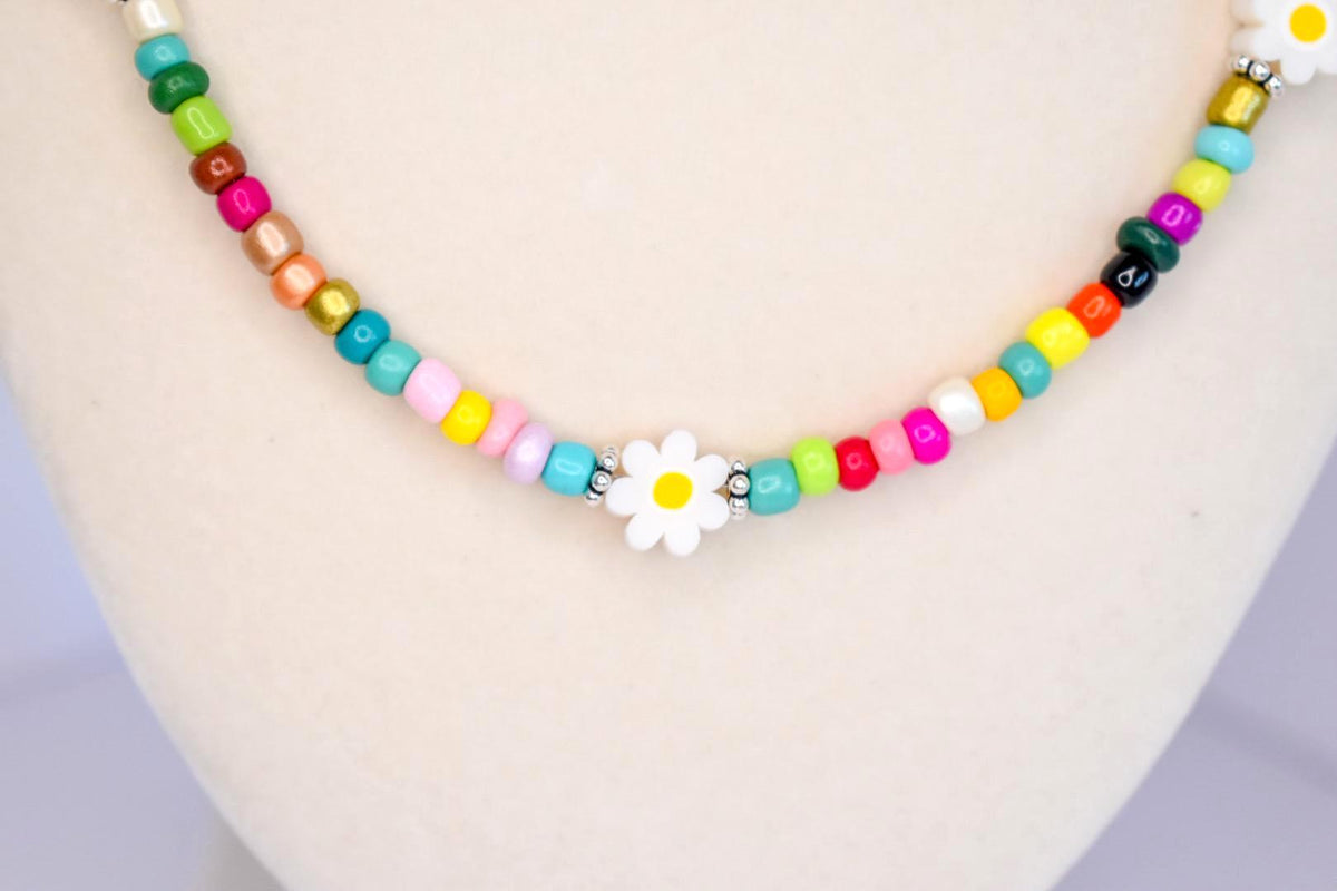 A beaded necklace featuring a variety of small, colorful round beads in shades of pink, blue, green, yellow, and metallic tones, accented with white daisy-shaped flower beads with yellow centers and small silver-toned spacer beads.
