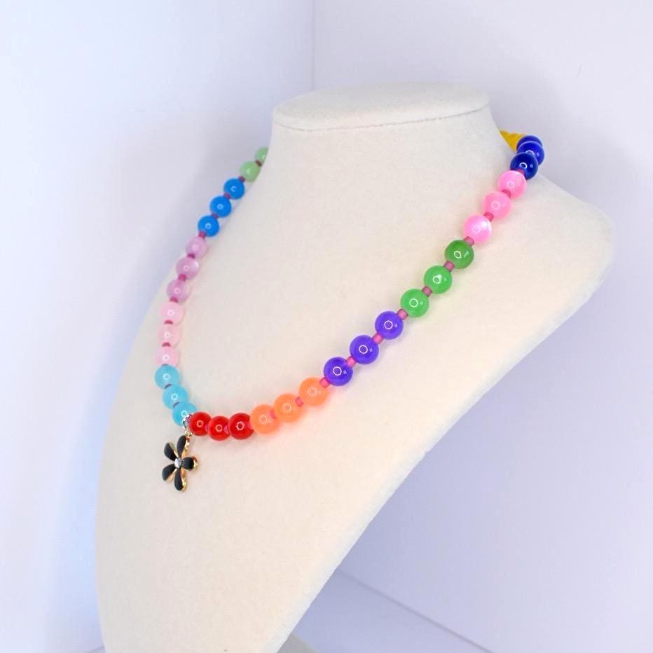 A colorful beaded necklace with a flower pendant. The necklace features a gradient of beads in various colors, including blue, pink, green, purple, orange, and red. The pendant is a black flower with gold detailing. The necklace is displayed on a white bust.
