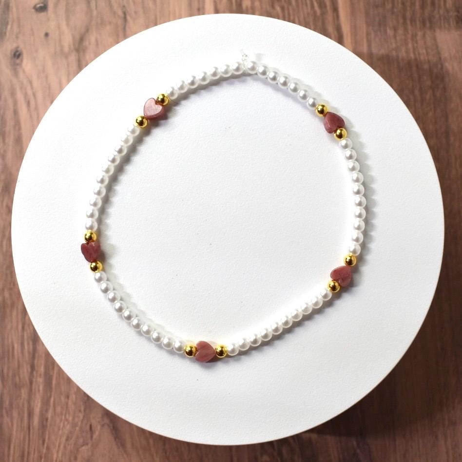 A delicate bracelet featuring small, white faux pearls interspersed with pink heart-shaped beads and gold-colored accents. The bracelet is displayed on a white surface, showcasing its elegant design. A charming piece of jewelry.