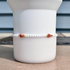 A delicate bracelet featuring small white pearls, interspersed with gold-colored beads and heart-shaped pink gemstones. The bracelet is displayed against a white background, showcasing its elegant design and craftsmanship.
