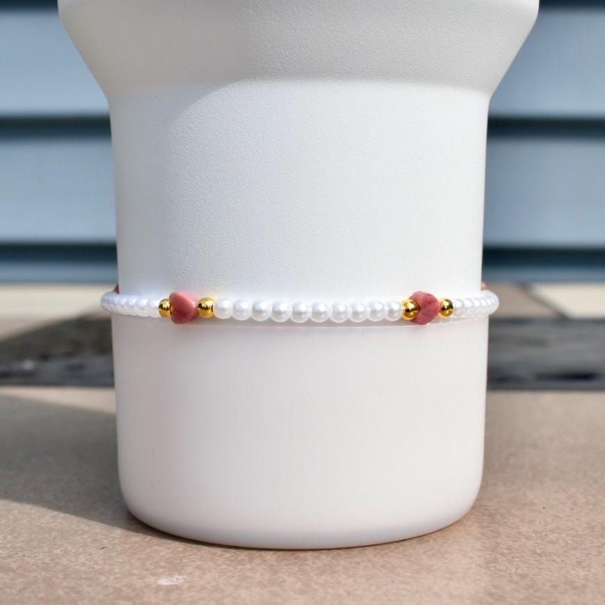 A delicate bracelet featuring small white pearls, interspersed with gold-colored beads and heart-shaped pink gemstones. The bracelet is displayed against a white background, showcasing its elegant design and craftsmanship.