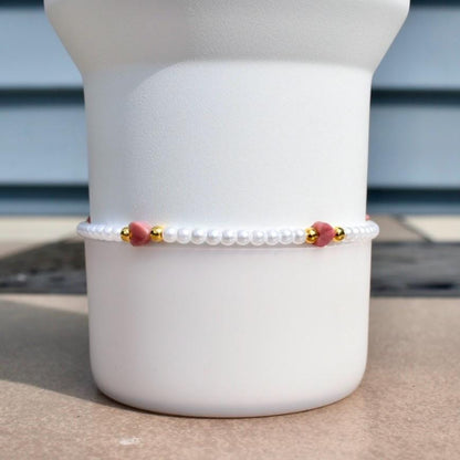 A delicate bracelet featuring small white pearls, interspersed with gold-colored beads and heart-shaped pink gemstones. The bracelet is displayed against a white background, showcasing its elegant design and craftsmanship.