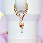 Gold-toned chain with a heart-shaped pendant. The pendant features a pink rose design on a cream background, with a small, sparkling charm dangling below. The chain is attached to a white object.
