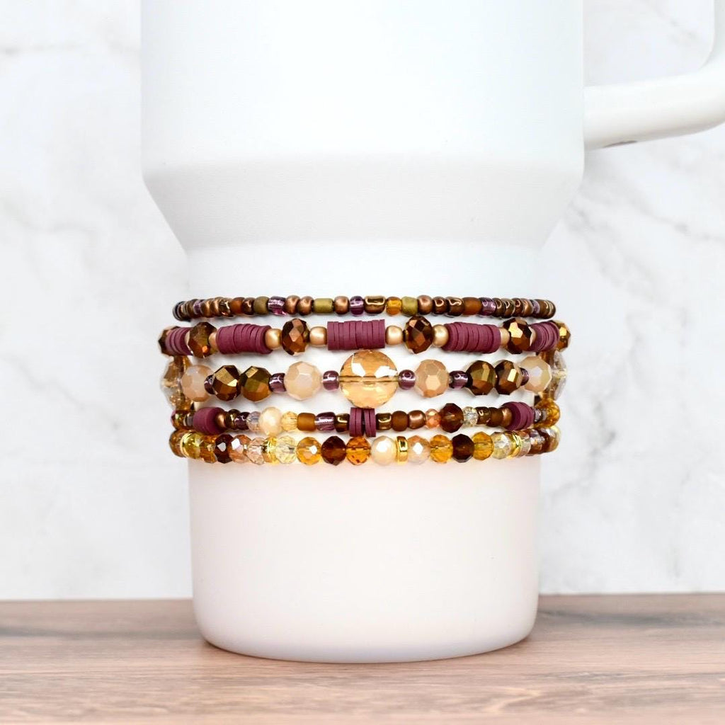 A stack of beaded bracelets in shades of brown, gold, and burgundy, resting on a white mug. The bracelets feature various bead sizes and textures, creating a layered effect. The background is a light marble pattern.