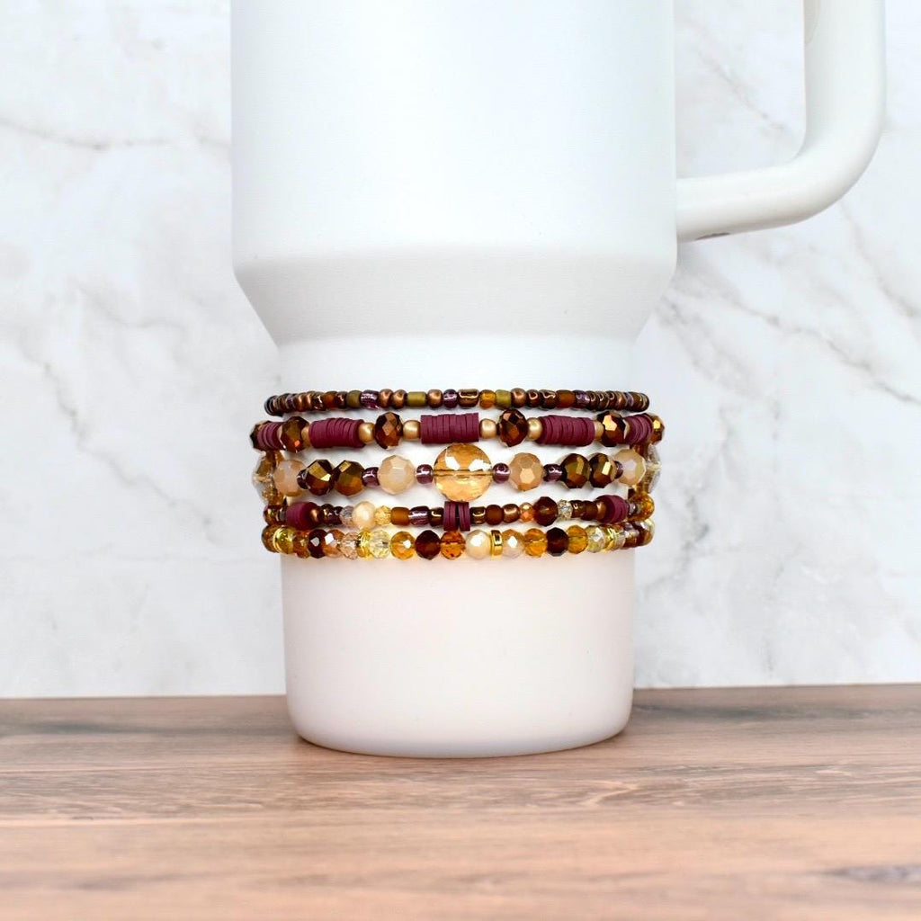 A stack of beaded bracelets in shades of burgundy, gold, and brown adorns a white tumbler. The bracelets feature various bead sizes and textures, creating a layered effect. The background is a white marble pattern.