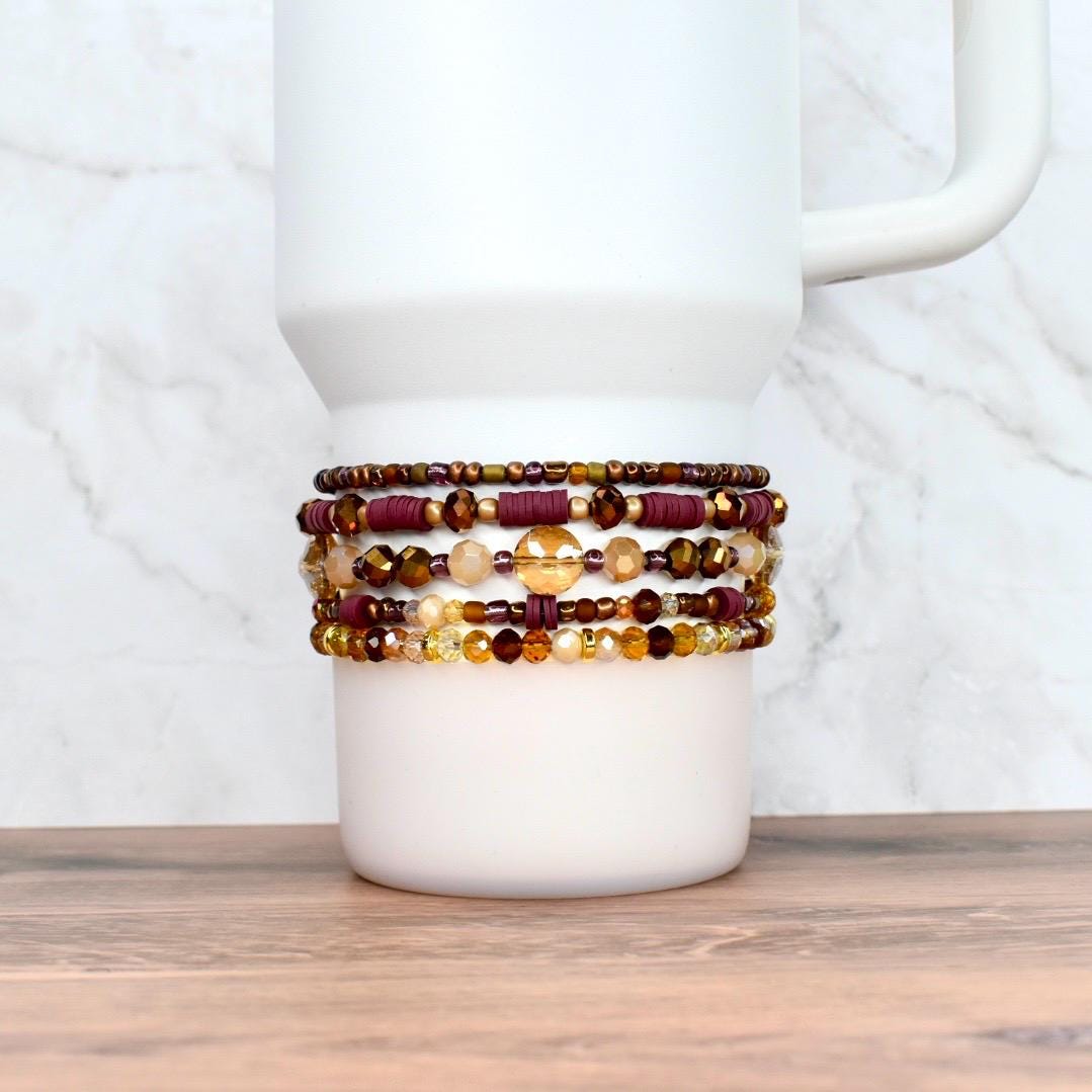 A stack of beaded bracelets in shades of burgundy, gold, and brown adorns a white tumbler. The bracelets feature various bead sizes and textures, creating a layered effect. The background is a white marble pattern.