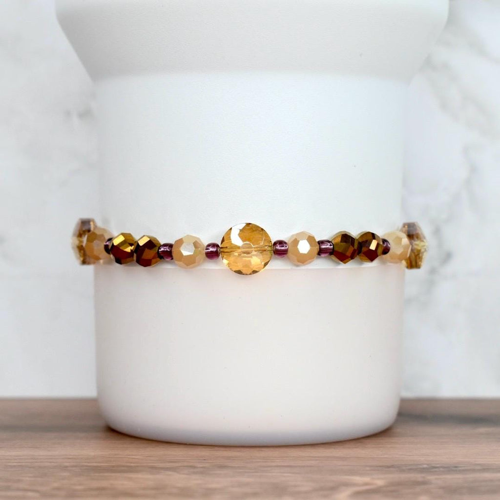 A beaded bracelet featuring a mix of amber, gold, and dark brown faceted beads. The bracelet includes a large, round, gold-colored bead as a focal point, with smaller purple beads interspersed throughout. The bracelet is displayed against a white background.