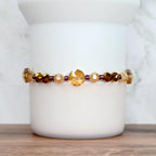 A beaded bracelet featuring a mix of amber, gold, and dark brown faceted beads. The bracelet includes a large, round, gold-colored bead as a focal point, with smaller purple beads interspersed throughout. The bracelet is displayed against a white background.