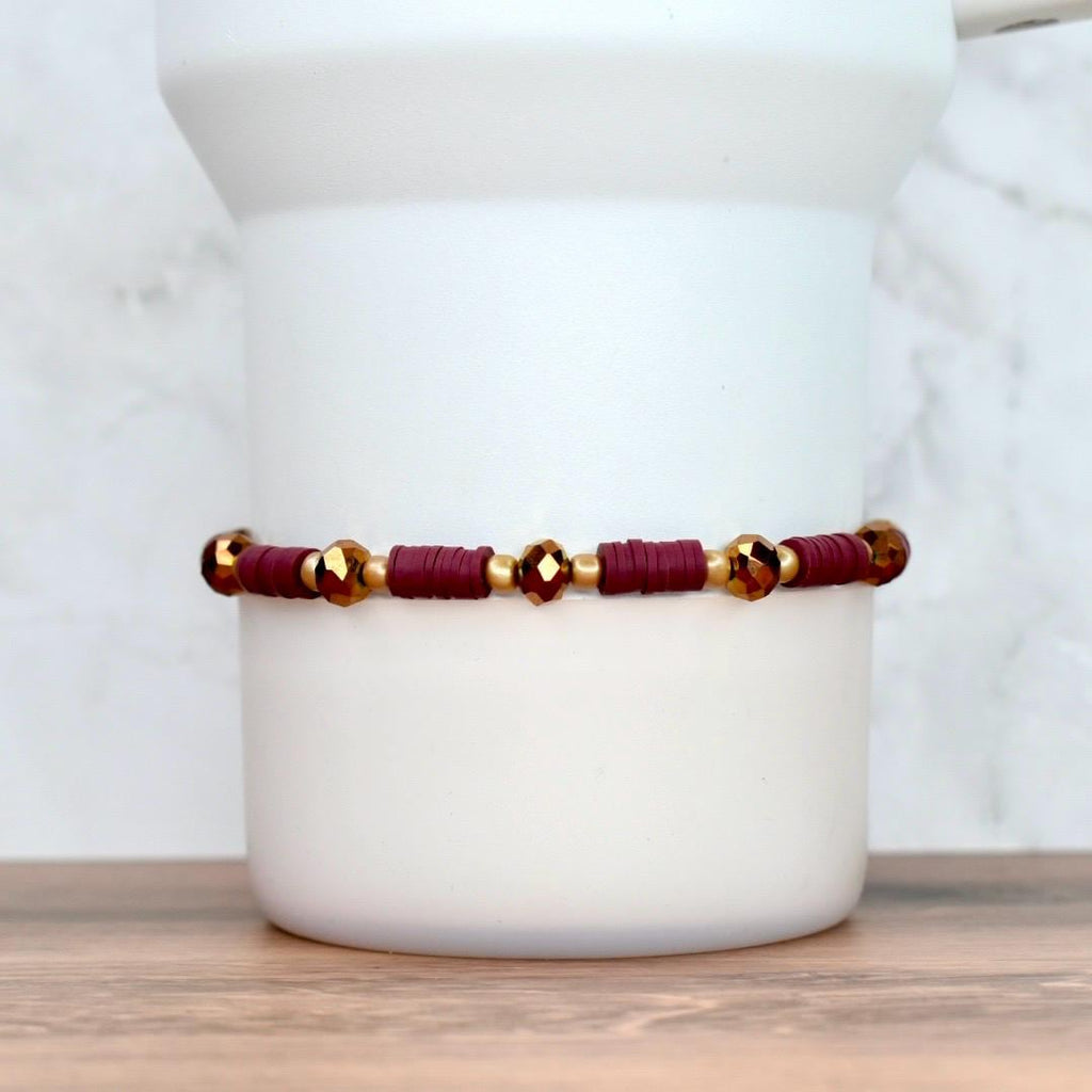 A burgundy and gold beaded bracelet. The bracelet features small, flat burgundy beads interspersed with gold-colored beads and faceted gold-colored beads. The bracelet is displayed against a white background.