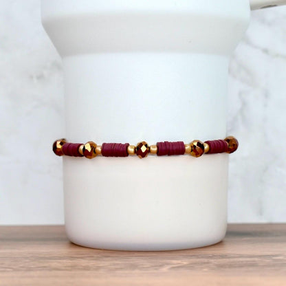 A burgundy and gold beaded bracelet. The bracelet features small, flat burgundy beads interspersed with gold-colored beads and faceted gold-colored beads. The bracelet is displayed against a white background.