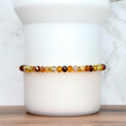 A beaded bracelet with a mix of amber, brown, and gold-colored faceted beads. The bracelet is strung on a gold-colored elastic cord. The beads are arranged in a pattern of varying sizes and shades, creating a warm, earthy aesthetic. The bracelet is displayed on a white cylindrical object.