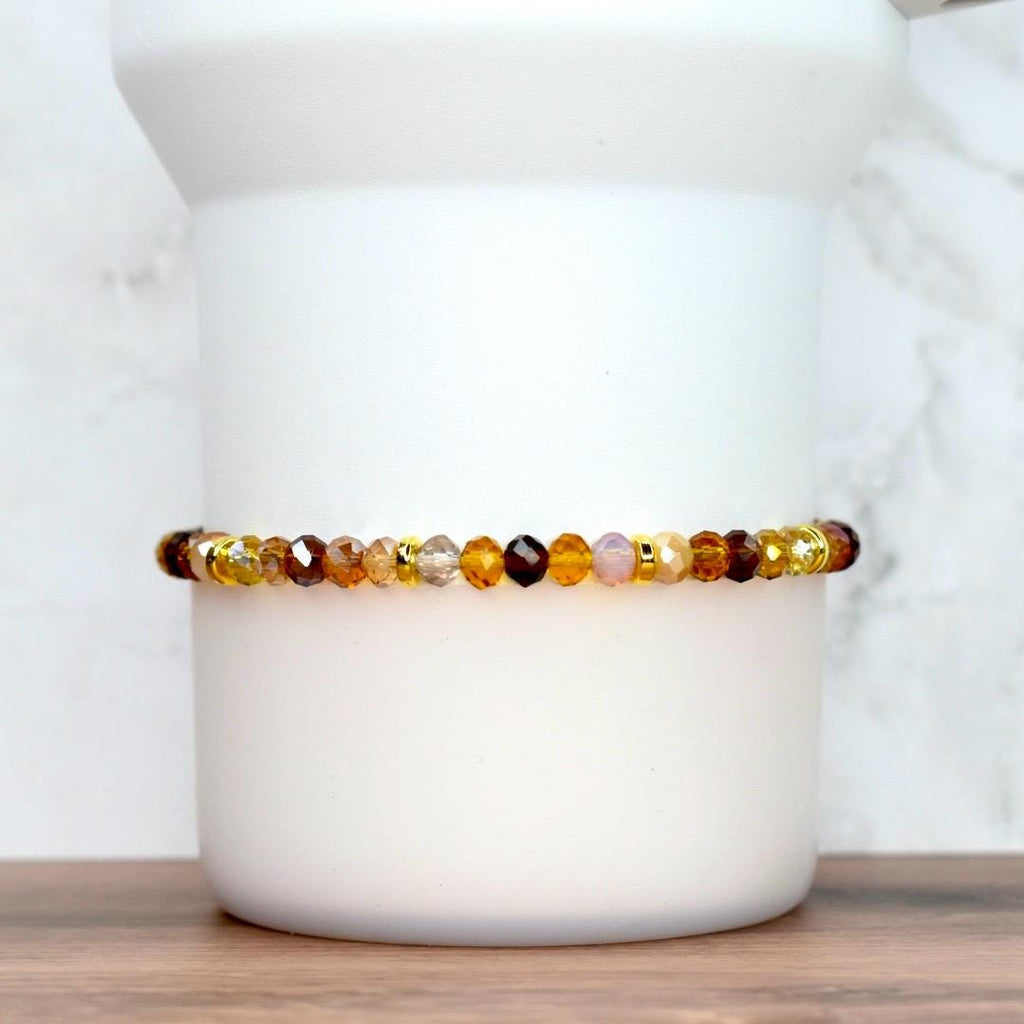 A beaded bracelet with a mix of amber, brown, and gold-colored faceted beads. The bracelet is strung on a gold-colored elastic cord. The beads are arranged in a pattern of varying sizes and shades, creating a warm, earthy aesthetic. The bracelet is displayed on a white cylindrical object.