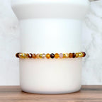 A beaded bracelet with a mix of amber, brown, and gold-colored faceted beads. The bracelet is strung on a gold-colored elastic cord. The beads are arranged in a pattern of varying sizes and shades, creating a warm, earthy aesthetic. The bracelet is displayed on a white cylindrical object.