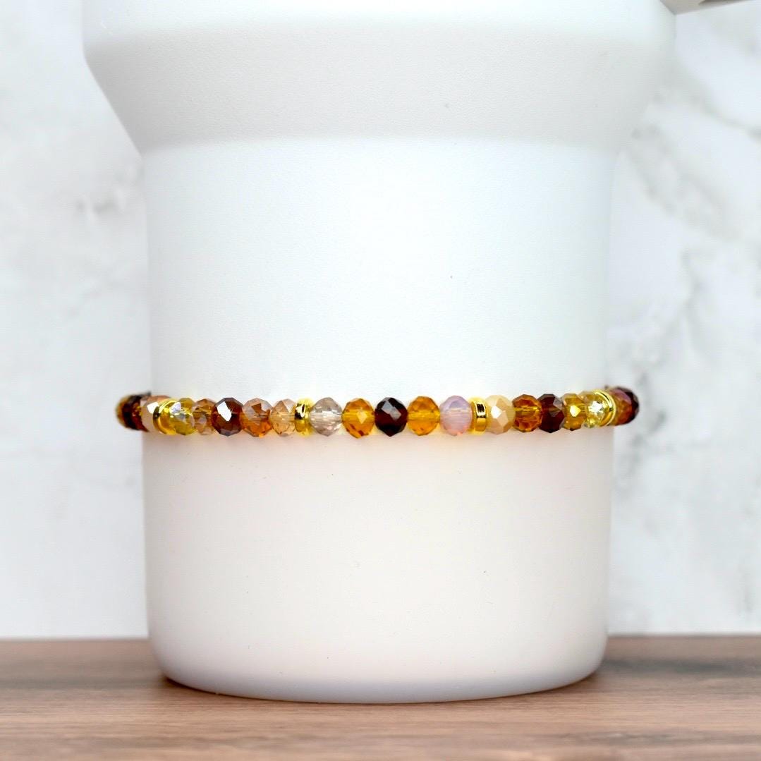 A beaded bracelet with a mix of amber, brown, and gold-colored faceted beads. The bracelet is strung on a gold-colored elastic cord. The beads are arranged in a pattern of varying sizes and shades, creating a warm, earthy aesthetic. The bracelet is displayed on a white cylindrical object.