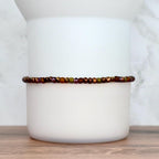A beaded bracelet with a mix of brown, purple, and green beads. The bracelet is resting on a white ceramic object. The beads are small and evenly spaced, creating a delicate and colorful accessory.